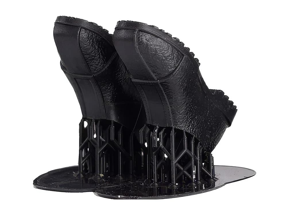 Zwei schwarze, glänzende Schuhsohlen aus ABS110, noch auf ihren gedruckten Stützstrukturen stehend.