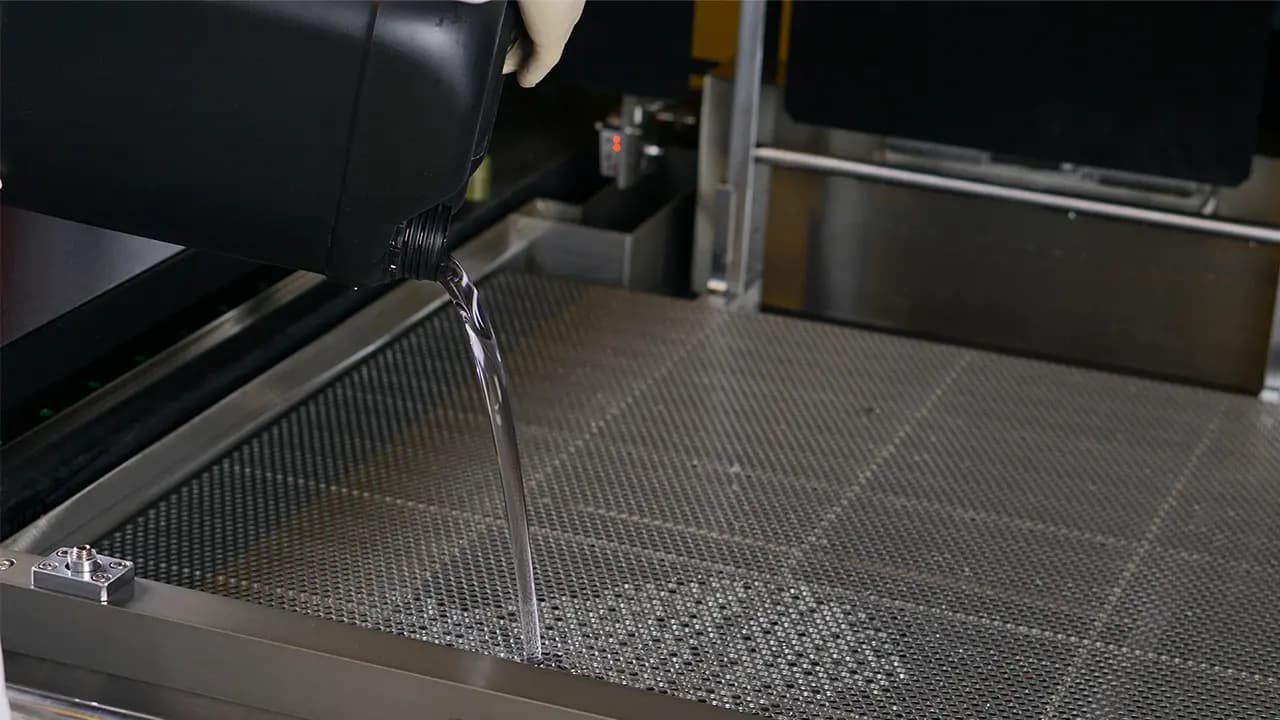 Eine Hand gießt Flüssigkeit aus einem schwarzen Behälter auf eine Metallgitterfläche.