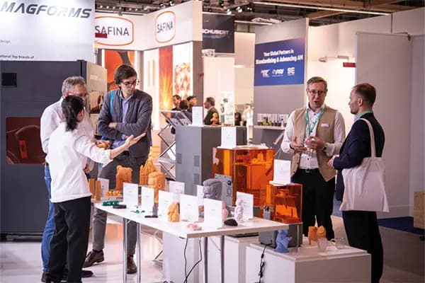 Menschen unterhalten sich an einem Messestand mit orangefarbenen 3D-Druckern und gedruckten Modellen auf Tischen.