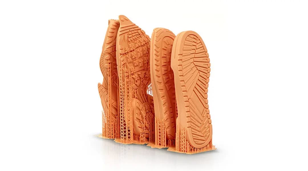 3D-gedruckte orangefarbene Schuhsohlen mit detaillierten Mustern und Stützstrukturen auf weißem Hintergrund.